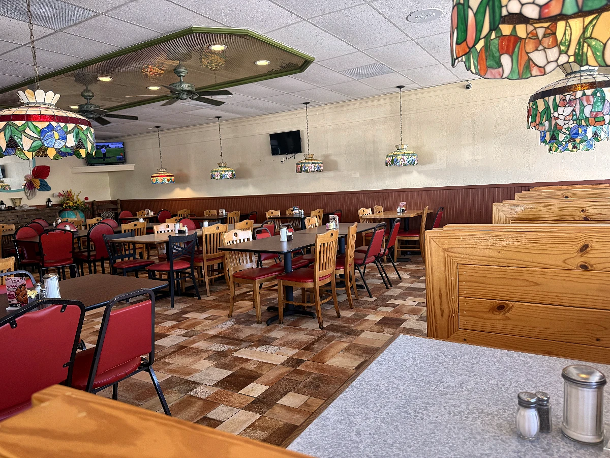 The main dining room: a full row of stained-glass pendants down the ceiling, ceiling fans, red vinyl chairs at laid tables, and pine booth backs along the right-hand side.