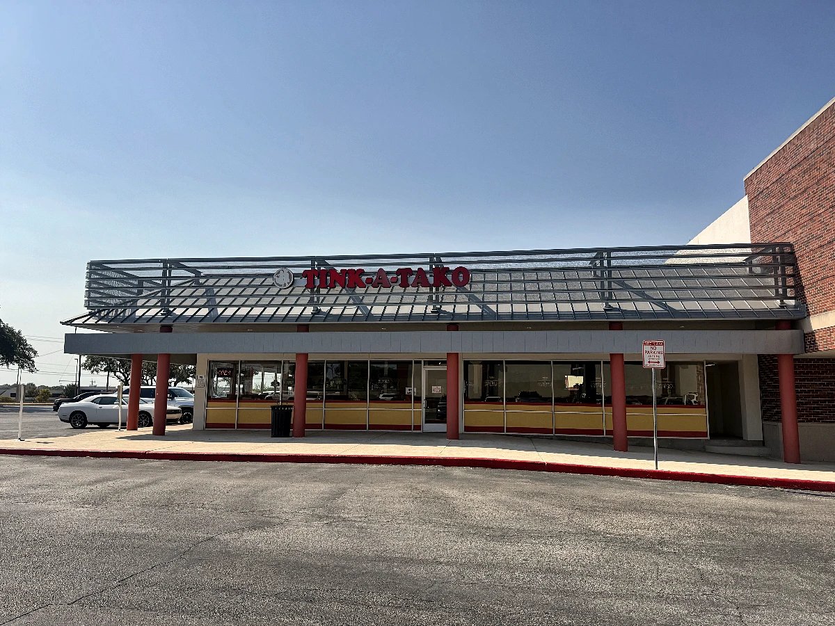 The storefront seen straight on: the Tink-A-Tako sign mounted on a steel trellis above the parapet, red columns along the walkway, a yellow and red band under the windows, and the parking lot in front.