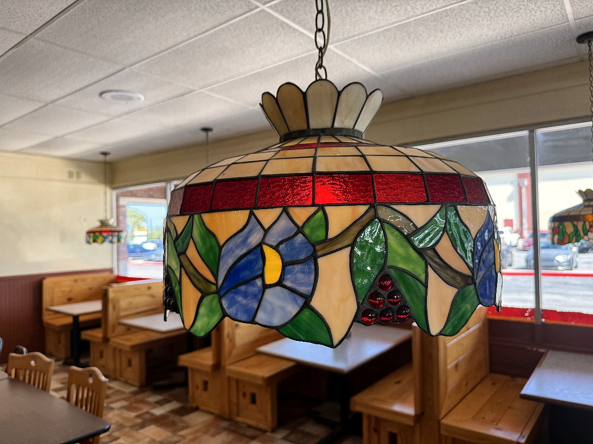 A stained-glass Tiffany pendant hanging low over the booths: blue morning-glory flowers and green leaves under a band of red glass, with pine booth backs receding behind it.
